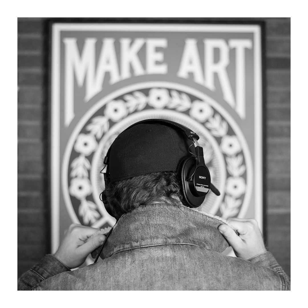 Black and white image of Joe Arden from behind, wearing headphones and a baseball cap in front of a Make Art poster